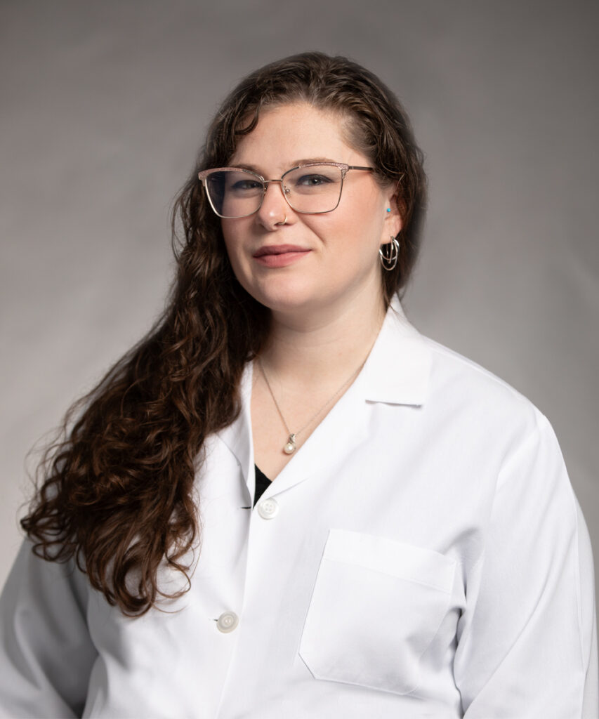 A woman with long, wavy brown hair and glasses is wearing a white lab coat and smiling slightly. The background is plain and neutral.