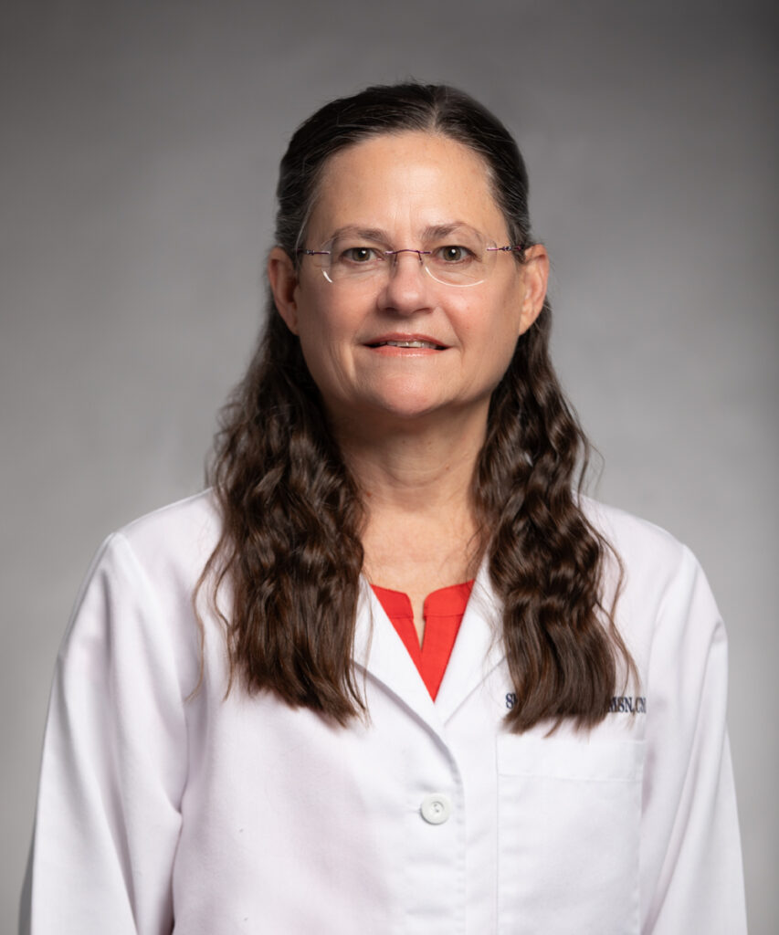 A middle-aged woman with long brown wavy hair and glasses, wearing a white lab coat over a red top, stands against a plain gray background, looking at the camera and smiling slightly.