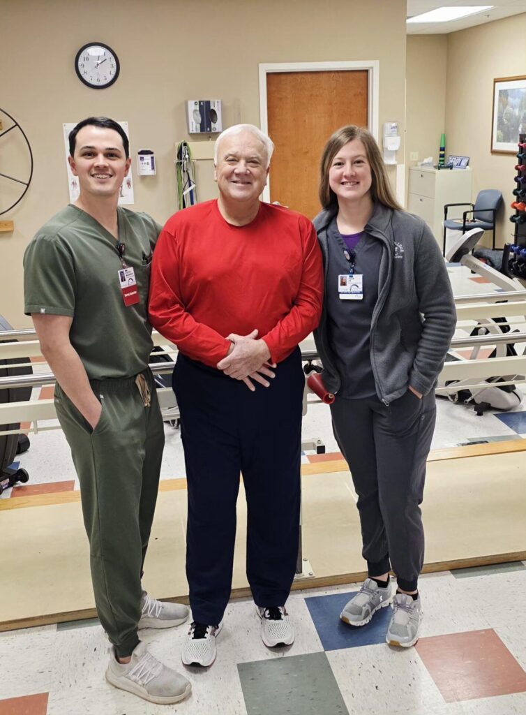 Three people stand smiling in a medical or physical therapy clinic. The man in the center wears a red shirt; the man on the left and woman on the right wear scrubs and name badges. Medical equipment is visible in the background.