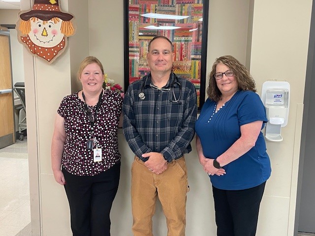 Three adults stand smiling in a hallway. A woman in a black patterned top is on the left, a man in a plaid shirt and khaki pants is in the center, and a woman in a blue top is on the right. Decorative art and a scarecrow hang on the wall.