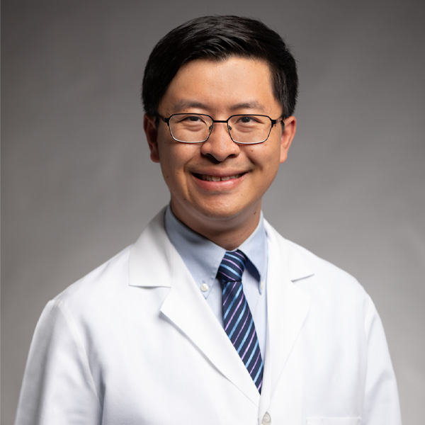 A man wearing glasses, a white lab coat, a light blue shirt, and a striped tie smiles at the camera against a plain, light gray background.