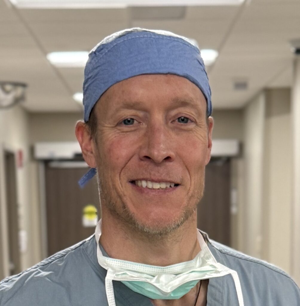 A smiling man in surgical scrubs and a blue cap stands in a hospital hallway, with a surgical mask hanging loosely around his neck.