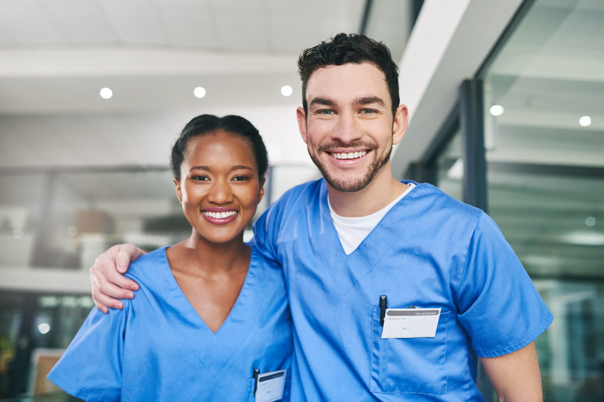 Two smiling healthcare professionals, one woman and one man, in blue scrubs with name badges, stand together indoors. They embody the supportive spirit of the Covenant Health nurse residency in a bright, modern setting.