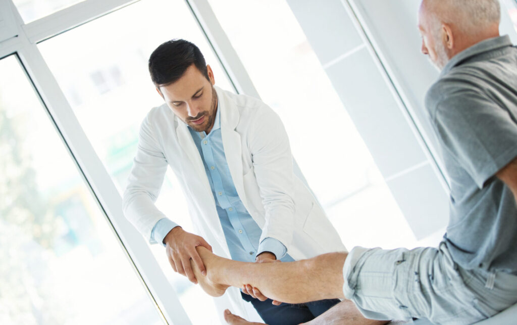 A doctor in a white coat examines the ankle and foot of an older man who is sitting on an exam table in a bright medical office.