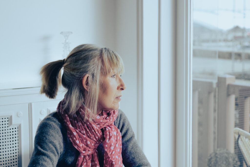 A woman with blonde hair in a ponytail and a pink scarf sits indoors, looking thoughtfully out a window. Soft daylight illuminates her face and the room around her.