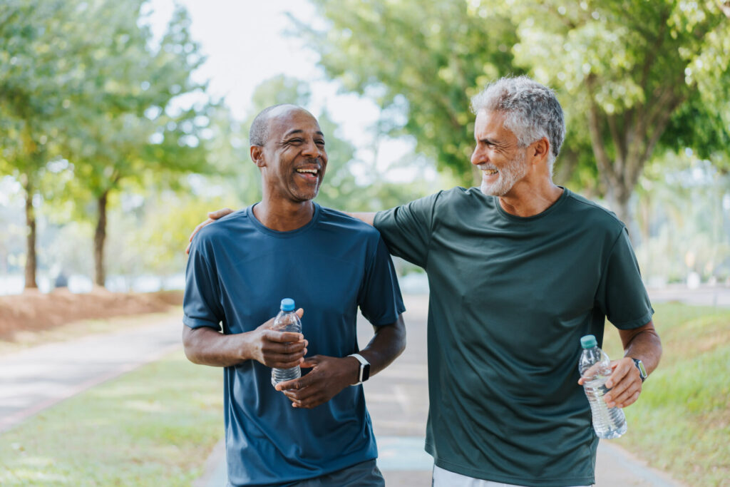 Two men walk outdoors on a sunny day, smiling and talking while holding water bottles.