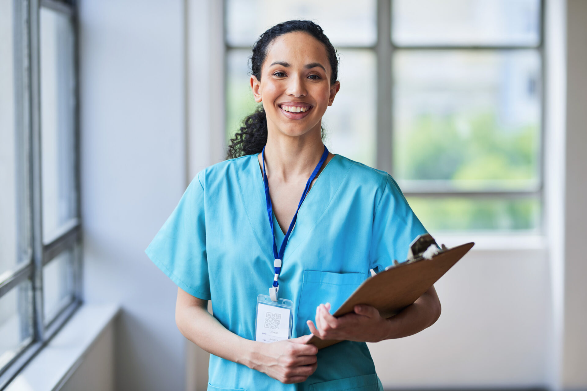 A Covenant Health nurse residency participant in light blue scrubs and a lanyard smiles while holding a clipboard in a brightly lit hallway with large windows in the background.