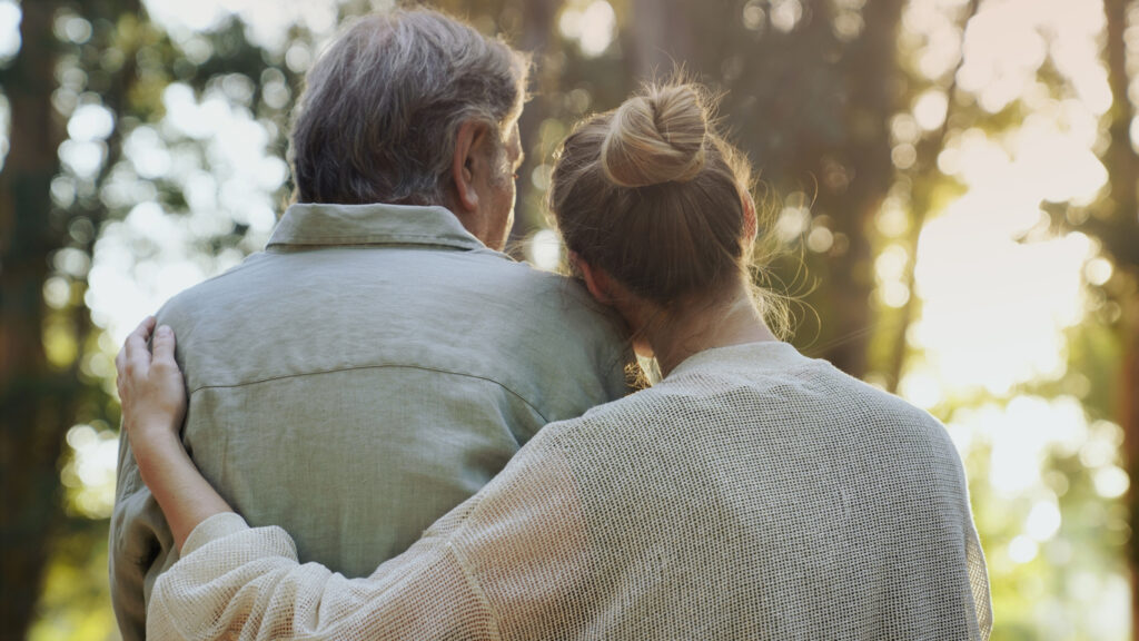 A woman with her arm around an older man, both seen from behind, stand closely together outdoors, surrounded by trees and soft sunlight.