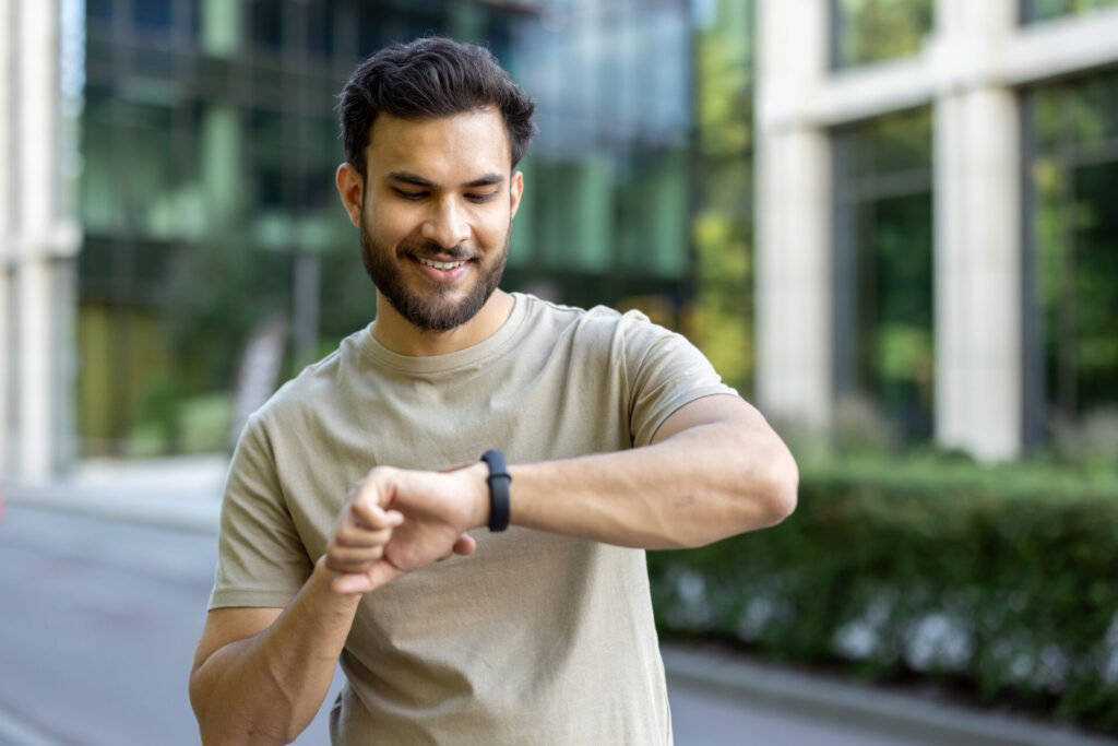 A smiling man looks at a smartwatch on his wrist while standing outdoors.