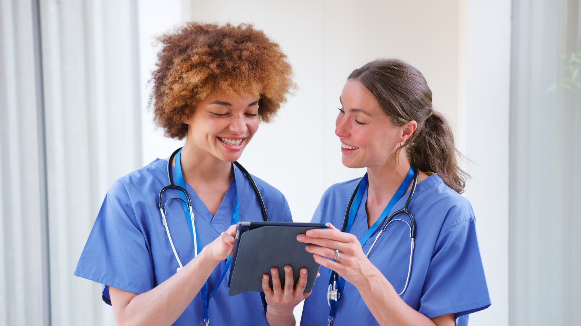 Two female healthcare professionals in blue scrubs and stethoscopes smile while looking at a tablet together, standing indoors in a bright medical setting—capturing the collaborative spirit of the Covenant Health nurse residency program.