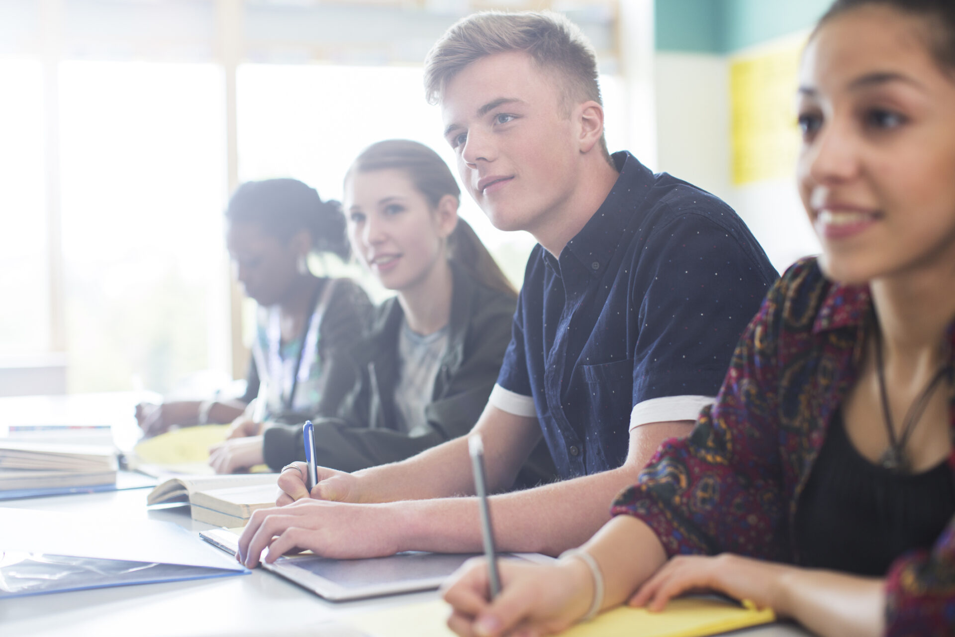 Four students sit at desks in a classroom during a High School Healthcare Seminar, writing in notebooks and looking attentive. Sunlight streams through the windows, creating a bright and focused learning environment.