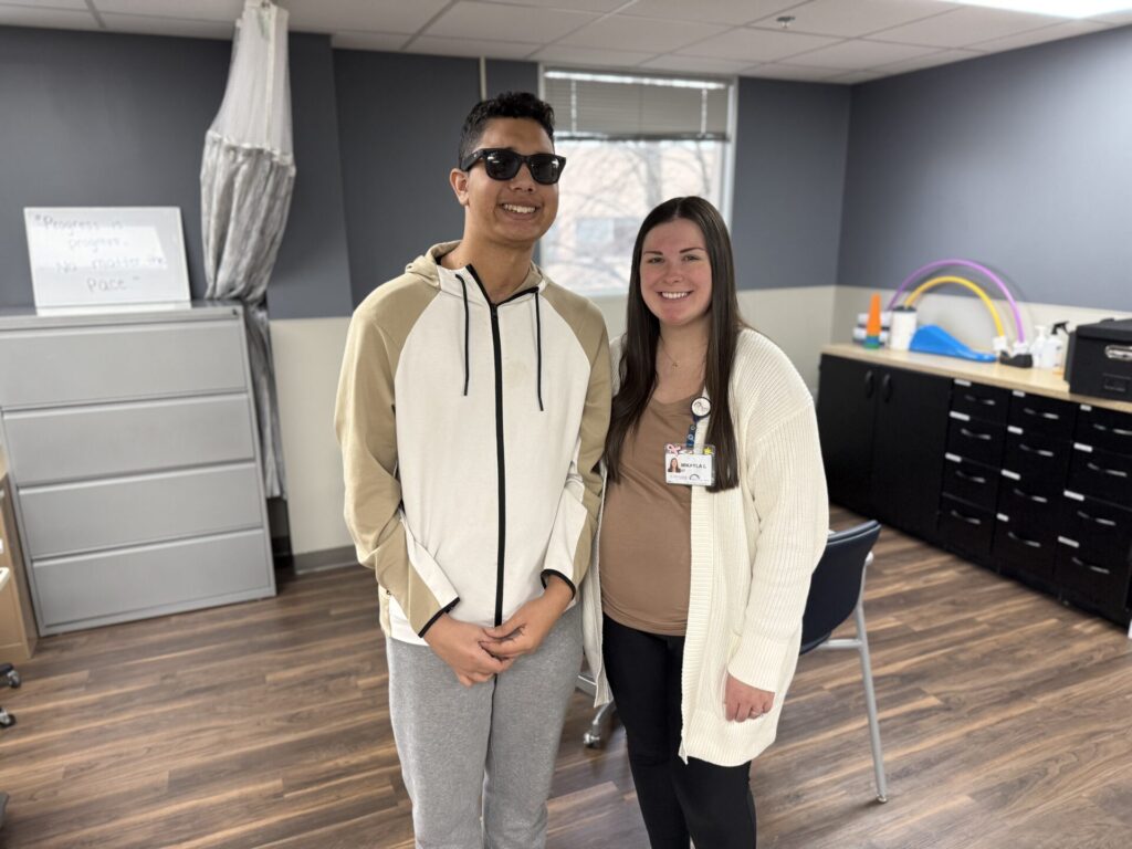 Two people stand smiling in a room with gray walls, wood flooring, and office furniture. One wears sunglasses and a hoodie; the other has a name badge and long brown hair. Bright therapy equipment is visible in the background.