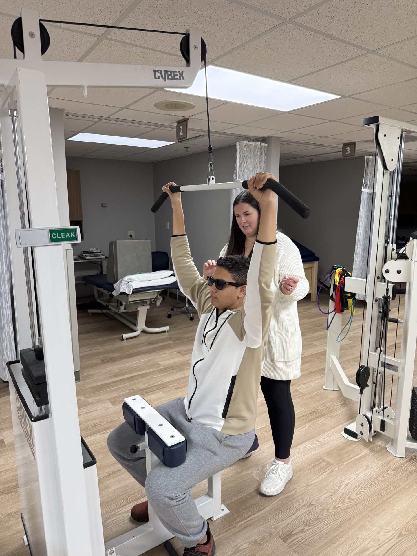 A person wearing sunglasses and a hoodie uses a lat pulldown machine while a woman in a white sweater stands behind, providing assistance in a physical therapy or gym setting.