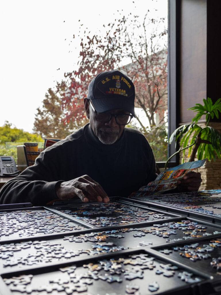 An older man wearing a U.S. Air Force Veteran hat works on a jigsaw puzzle by a window, holding a puzzle box for reference. Autumn trees and office items are visible in the background.