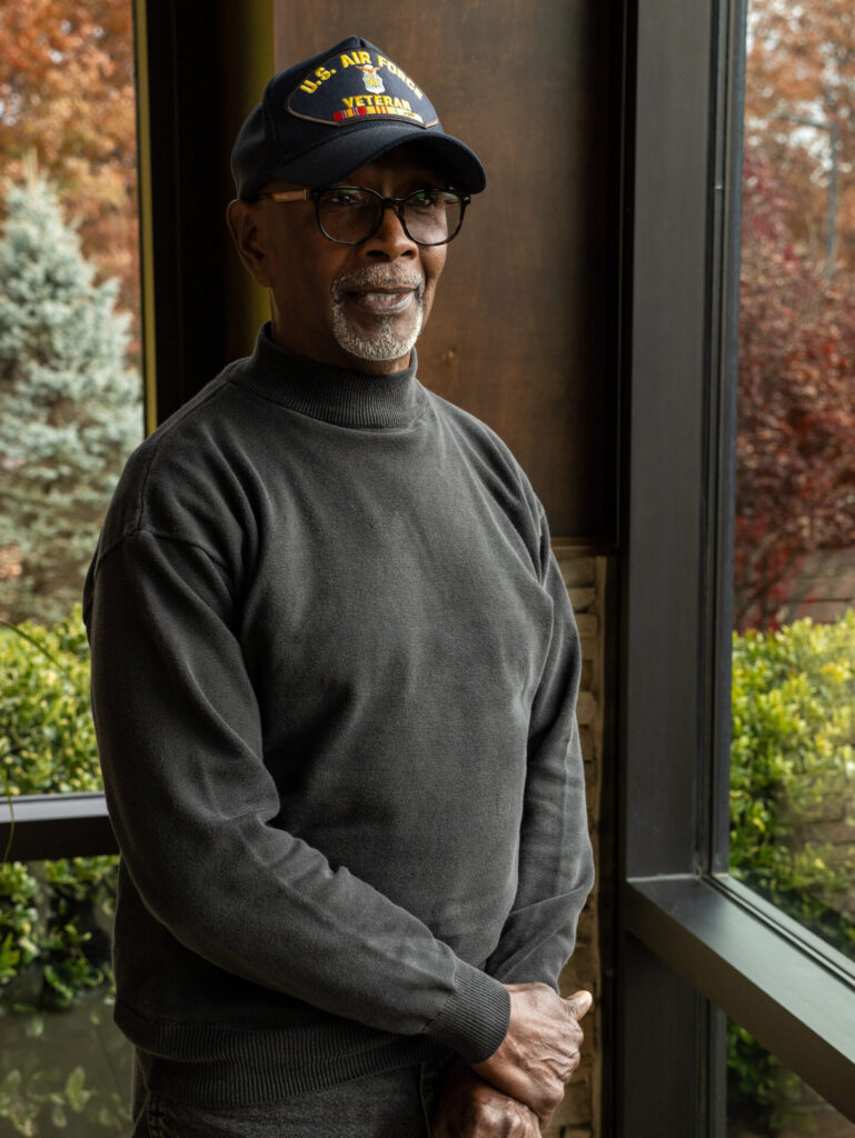 Edward Woodson, an older man wearing glasses, a dark cap labeled U.S. Air Force Veteran, and a dark sweater stands indoors by a window, with green bushes and autumn trees outside—reflecting on his journey through proton therapy.
