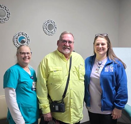 Three adults stand and smile indoors. Two women wear medical uniforms, one in teal scrubs and one in a blue RN jacket, and a man in a yellow shirt with a camera bag stands between them. Decorative mirrors hang on the wall behind.