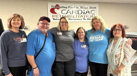 Six smiling adults stand together in front of a &ldquo;Cardiac Rehabilitation&rdquo; banner, celebrating Cardiac Rehab Week. Some wear blue &ldquo;Rehab Team&rdquo; shirts, and others wear badges, suggesting they are staff or participants.