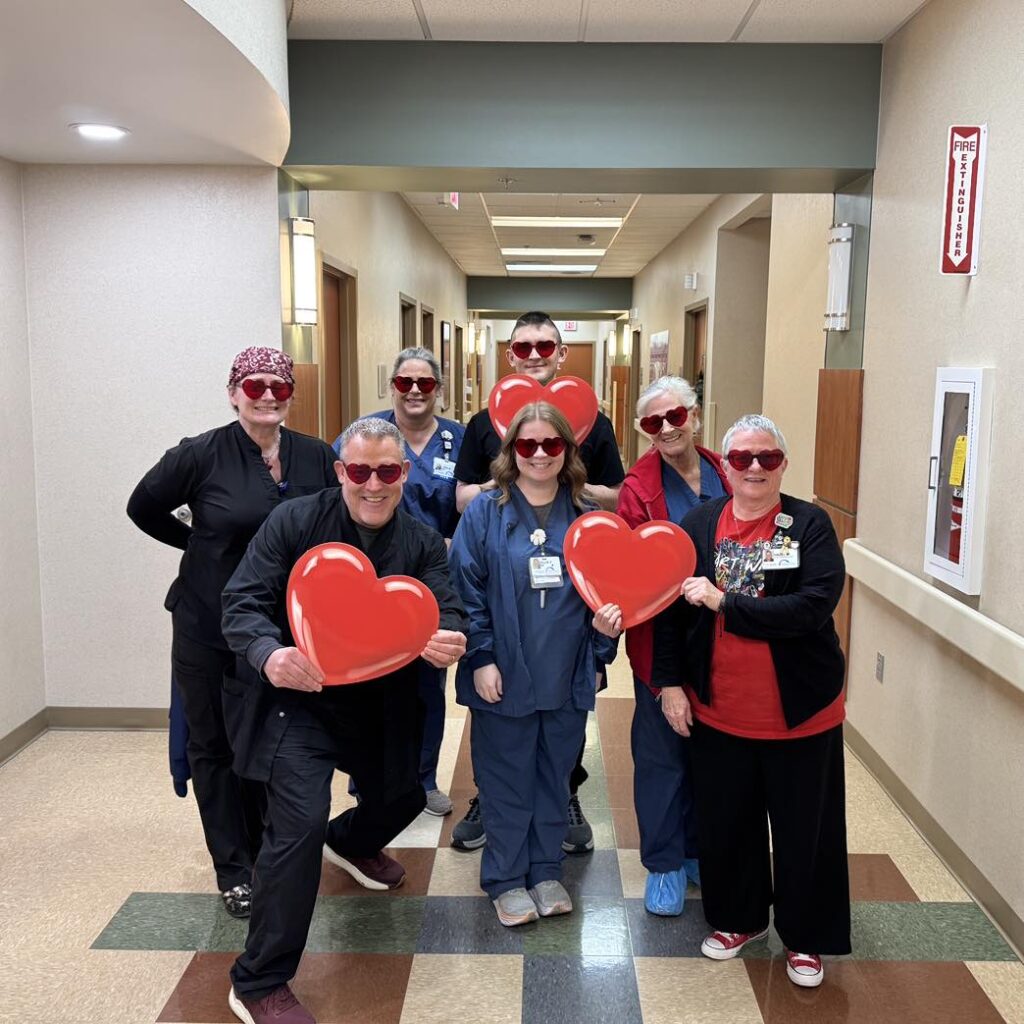 A group of healthcare workers in scrubs and uniforms stand in a hallway at Covenant Health Morristown-Hamblen, all wearing red heart-shaped sunglasses. Three hold large red heart props, celebrating Advanced Heart Care and their cheerful, united spirit.