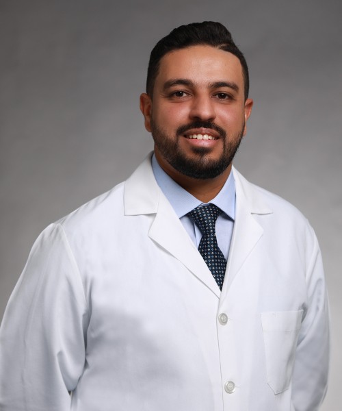 A man with short dark hair and a beard is smiling, wearing a white lab coat, light blue shirt, and a dark patterned tie, posed against a plain gray background.