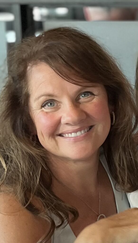 A woman with wavy brown hair and blue eyes smiles at the camera. She is wearing a white top, a necklace, and hoop earrings, with a blurred indoor background.
