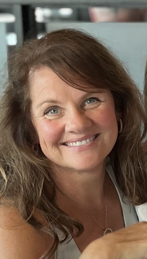 A woman with wavy brown hair and blue eyes smiles at the camera. She is wearing a white top, a necklace, and hoop earrings, with a blurred indoor background.