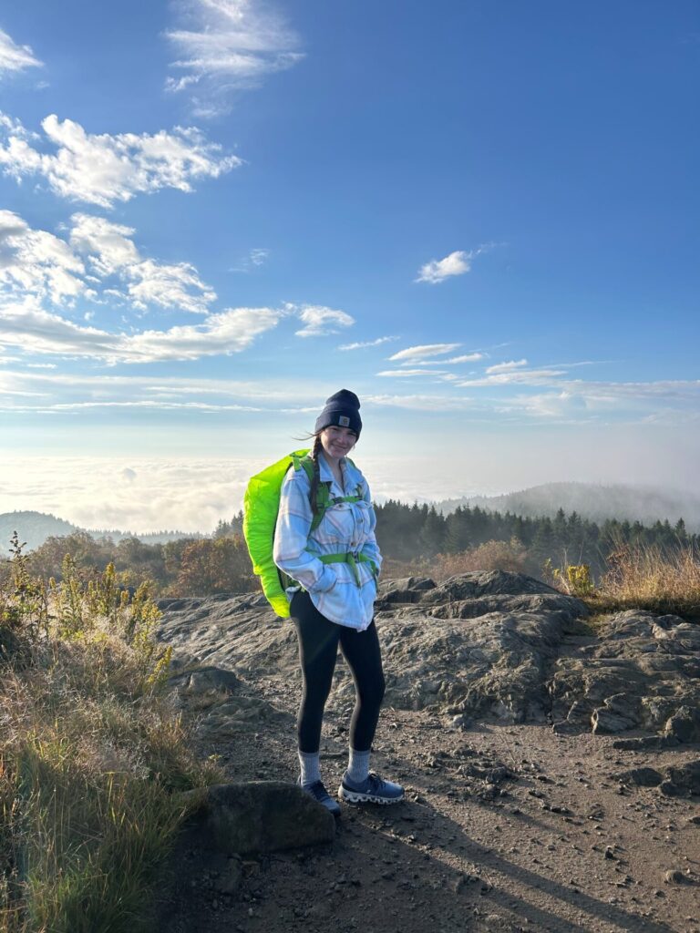 A person in a beanie, jacket, and leggings stands on a rocky hilltop with a bright green backpack, smiling at the camera. The background features a scenic view of trees, clouds, and blue sky.