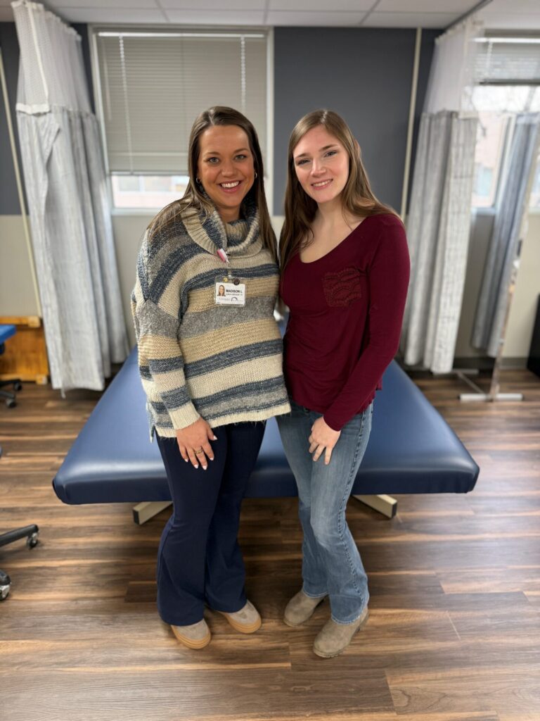 Two women stand smiling in front of a blue therapy table in a medical or physical therapy clinic. One wears a striped sweater and a name badge; the other wears a burgundy top and jeans. Curtains and windows are in the background.