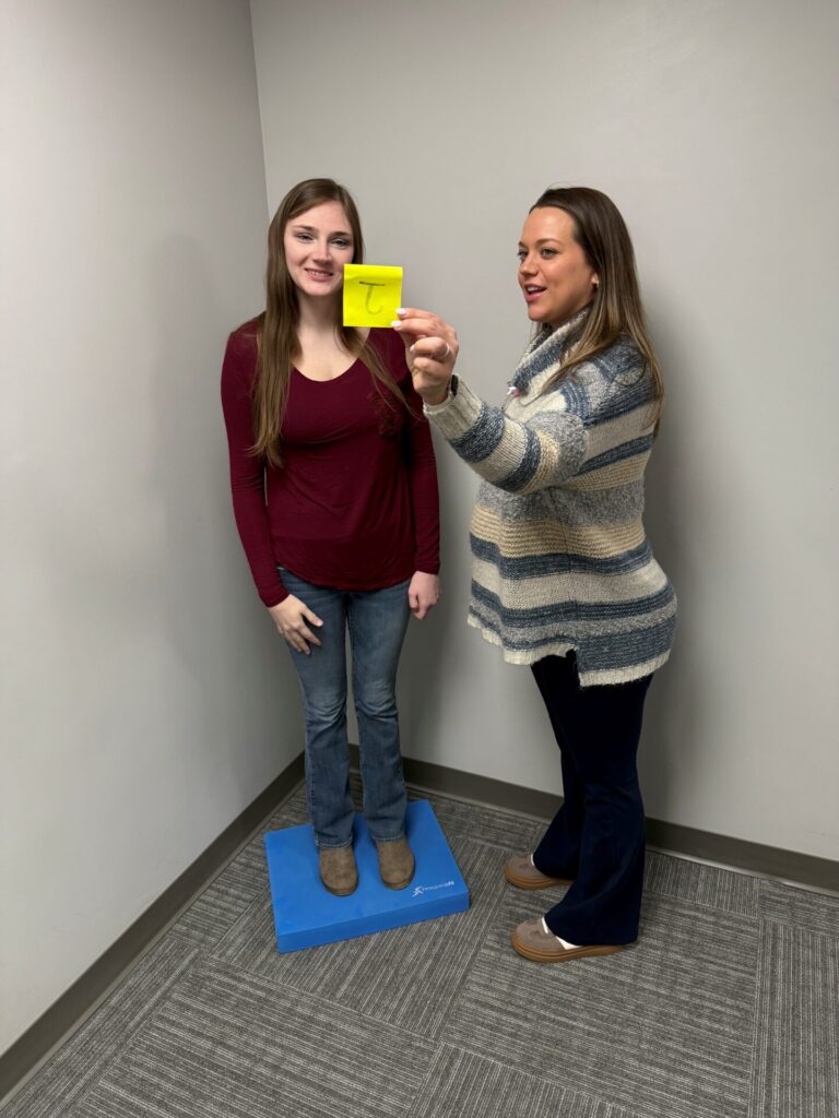 Two women stand in a corner; one is on a blue foam pad, smiling, while the other holds up a yellow card with the letter T written on it. They appear to be engaged in an activity or exercise.