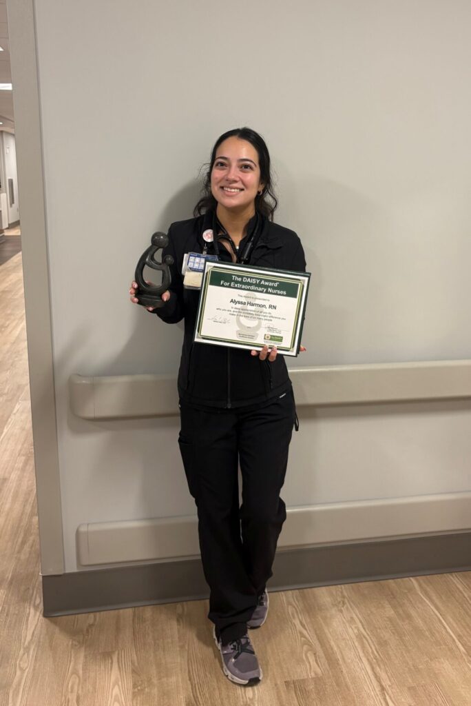 A smiling woman in black scrubs stands indoors holding a DAISY Award certificate in one hand and a sculpture in the other, posing against a hallway wall.