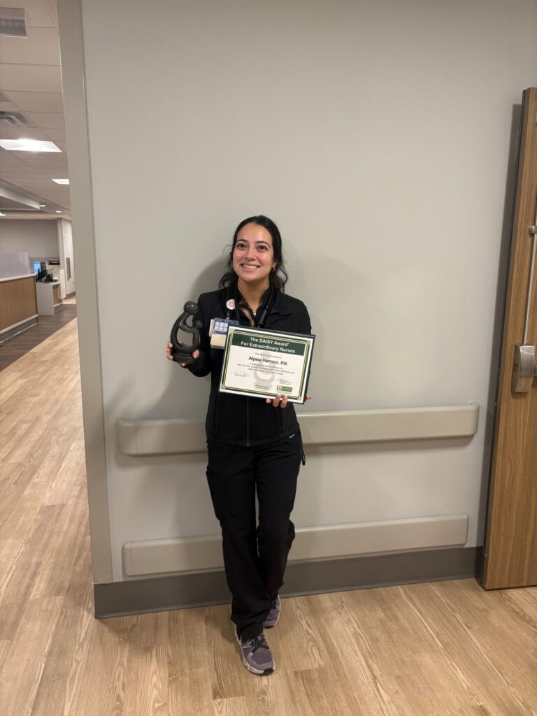 A woman in black scrubs stands in a hallway, smiling and holding an award certificate and a black trophy.