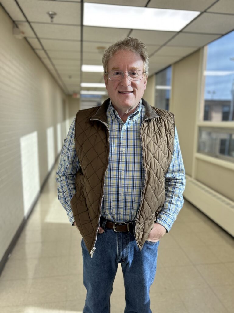 A man with gray hair and glasses stands in a bright hallway, smiling. He is wearing a brown quilted vest over a blue plaid shirt, blue jeans, and has one hand in his pocket. Sunlight streams in through large windows.
