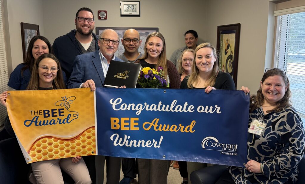 A group of smiling people stand indoors, holding a bouquet and two banners that read The BEE Award and Congratulate our Nancy Stanfield BEE Award Winner! Covenant Health Peninsulas.
