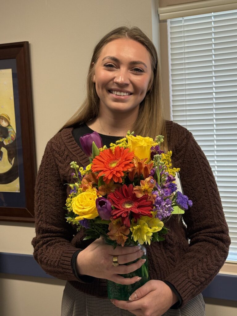 A smiling woman in a brown sweater holds a colorful bouquet of flowers, standing indoors near a window with blinds and a framed artwork on the wall behind her, celebrating her Nancy Stanfield BEE Award.