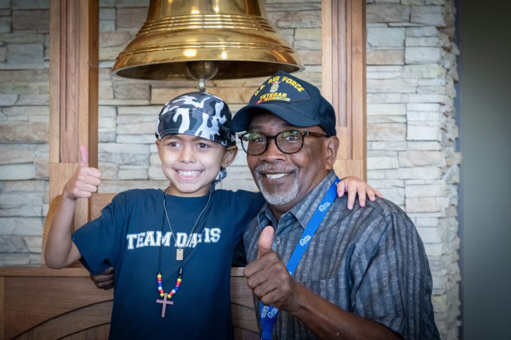 A smiling child wearing a camouflage cap and TEAM DAVIS shirt stands next to an older man in a U.S. Air Force Veteran hat; both give a thumbs-up in front of a large gold bell, celebrating after Edward Woodson proton therapy.