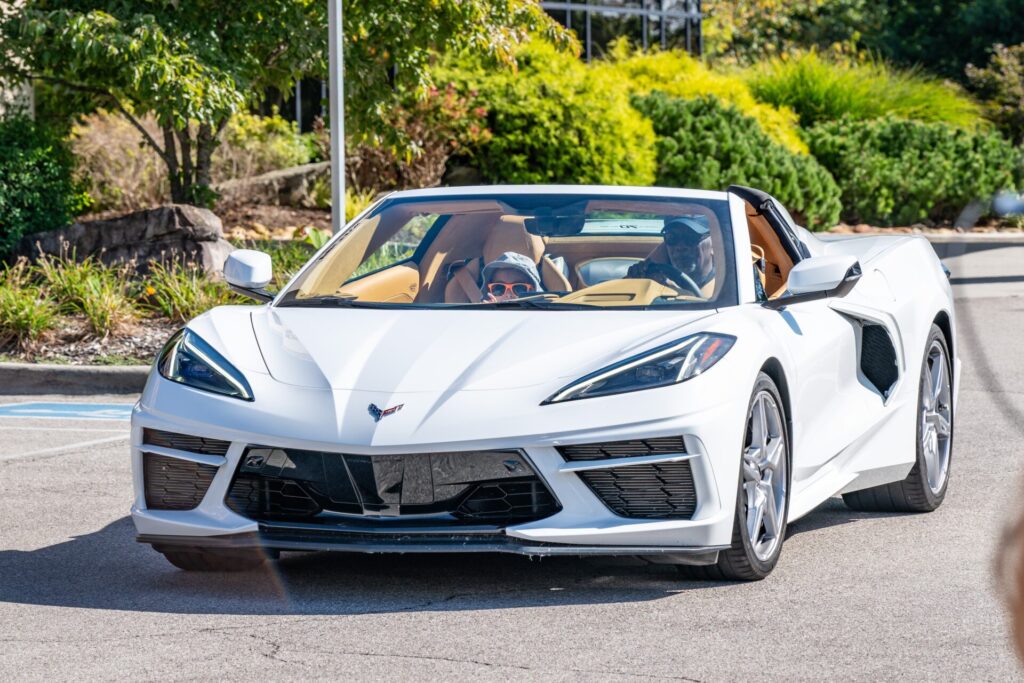 A white convertible sports car with two people inside drives on a sunny day. The driver, wearing sunglasses, chats about Edward Woodson and proton therapy as green bushes and trees line the background.