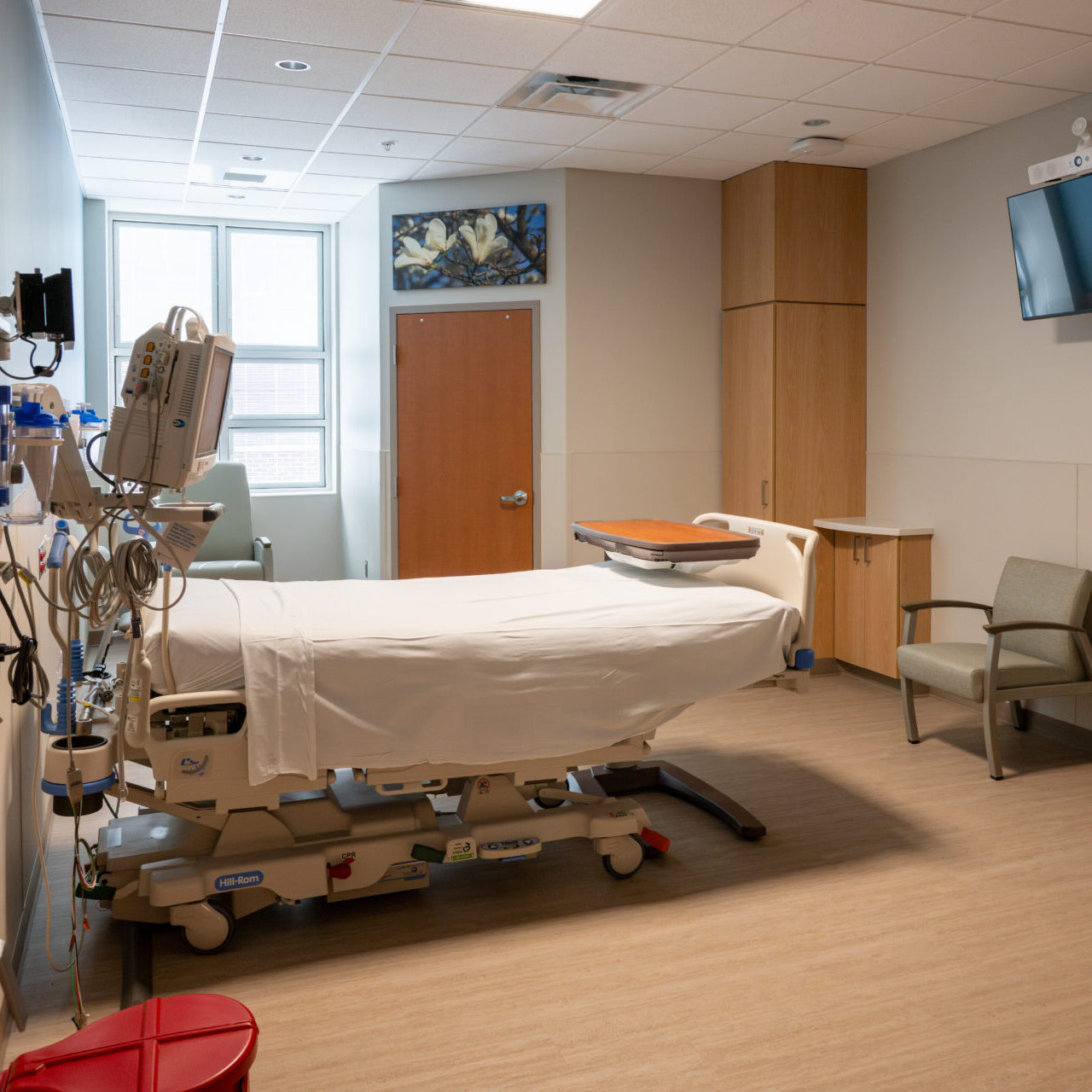 A modern hospital room with a single empty hospital bed, a gray chair, medical equipment on the wall, a flat-screen TV, wood cabinets, and a window letting in natural light.
