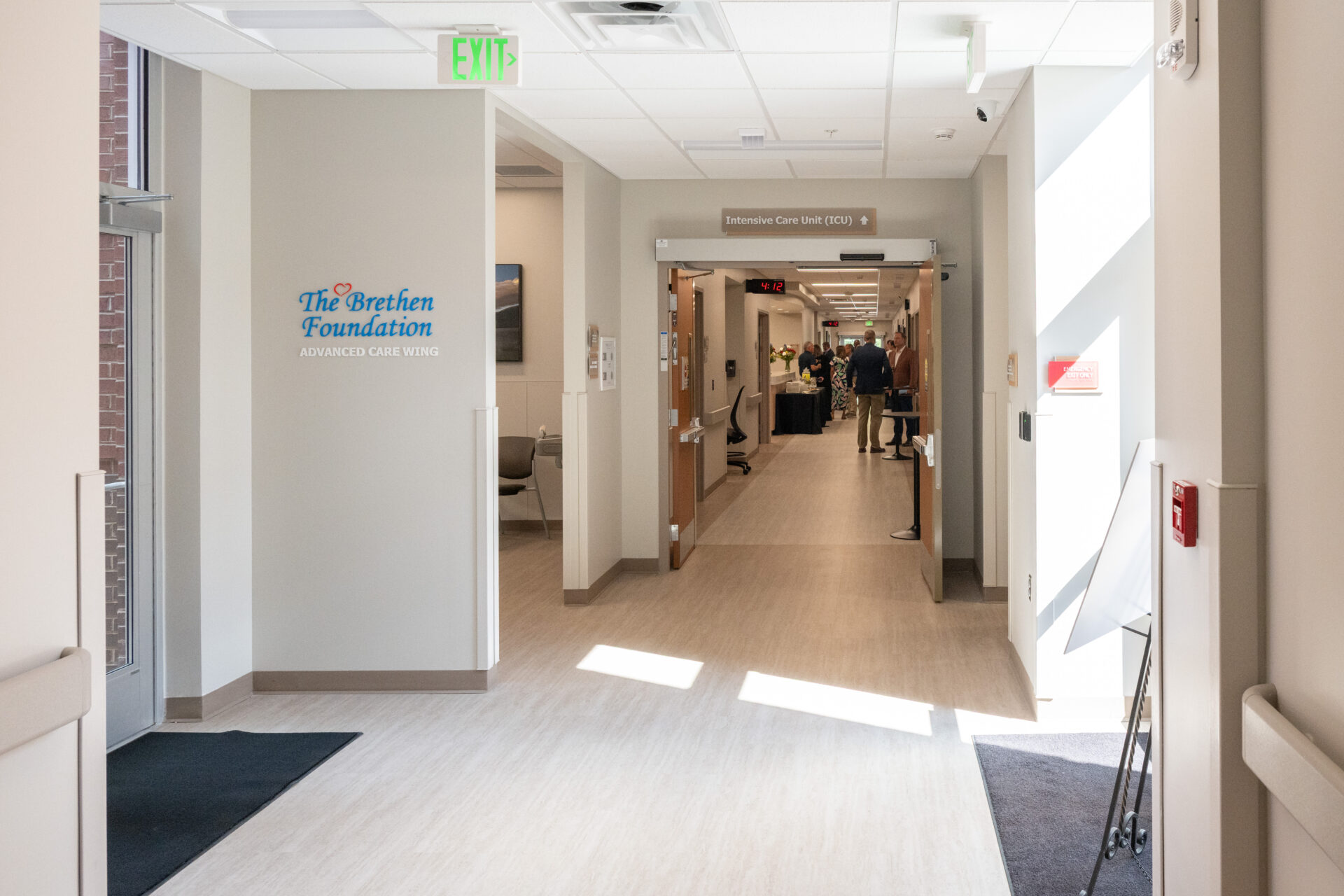 Bright hospital hallway with The Brethen Foundation Advanced Care Wing sign on the left wall, leading to the Intensive Care Unit (ICU); people are visible in the distance near the end of the corridor.