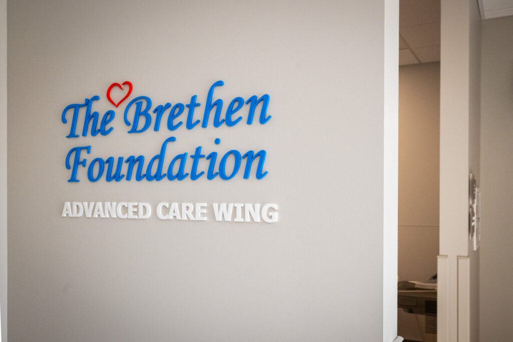 Wall sign reading The Brethen Foundation ADVANCED CARE WING in blue and white letters, with a red heart above the B. A doorway and part of an office interior are visible to the right.