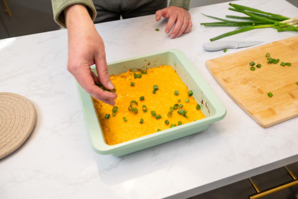 A person sprinkles freshly chopped green onions into a pan of buffalo chicken dip.