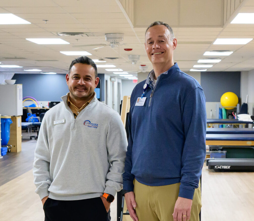 Two men standing and smiling in a bright, spacious fitness or therapy center with exercise equipment and large yellow fitness balls in the background. Both are wearing name tags and business casual attire.