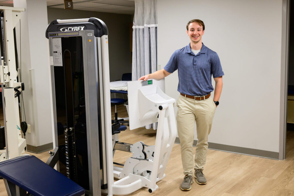 A man in a blue polo shirt and khaki pants stands smiling next to a seated leg press machine in a bright, modern gym or physical therapy room.