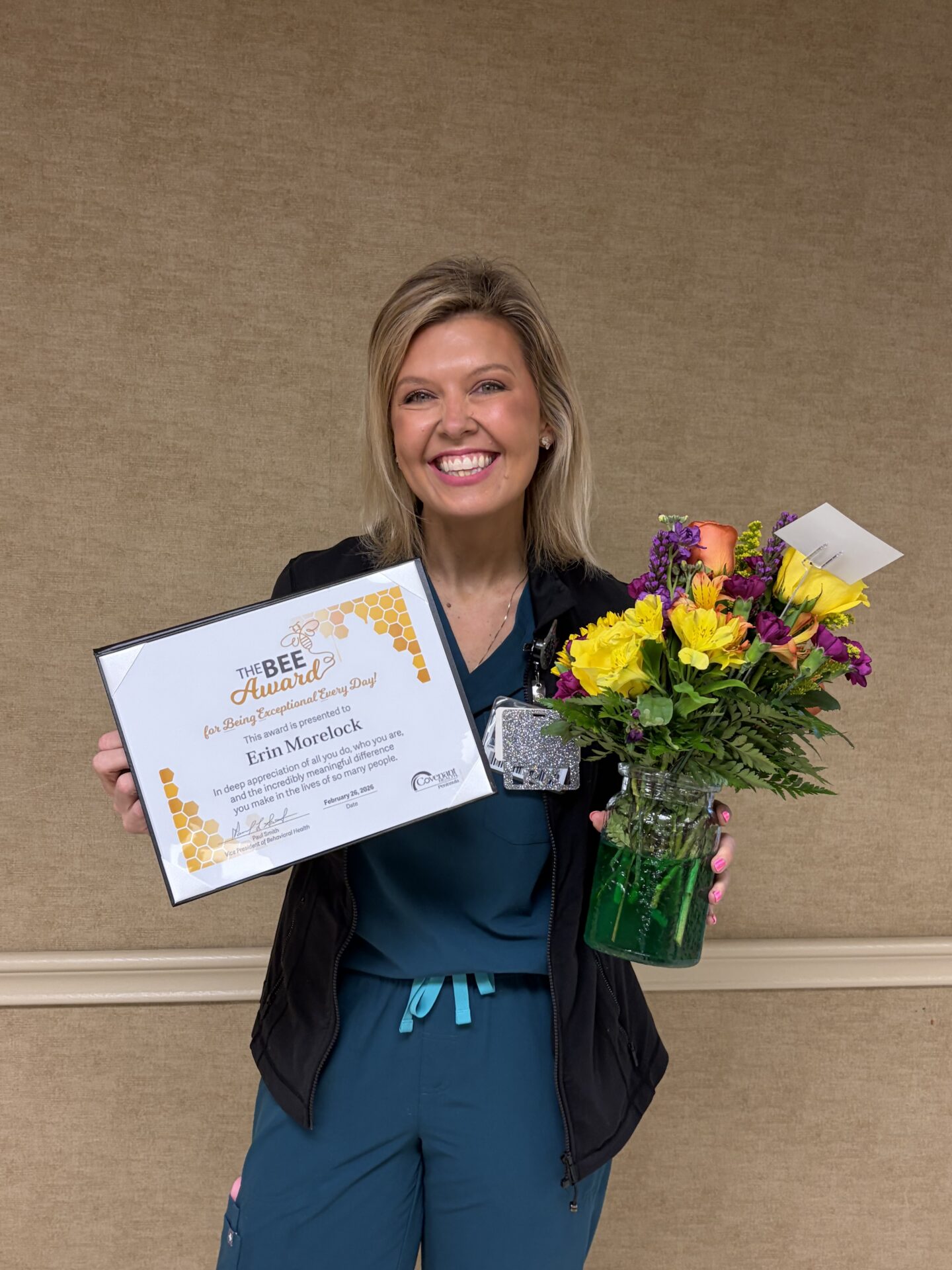 A smiling woman in scrubs holds a bouquet of colorful flowers and a framed award certificate, celebrating as Cassie Reed receives the BEE Award for Compassionate Care, standing against a tan wall.