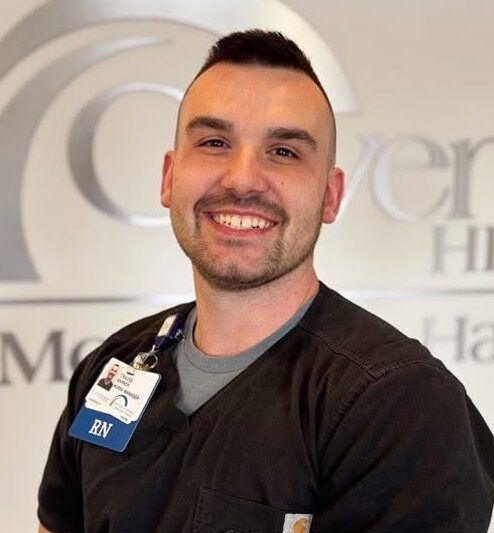 A smiling nurse in black scrubs and an RN badge stands in front of a wall with the words Genera Health, representing Advanced Heart Care at Covenant Health Morristown-Hamblen.