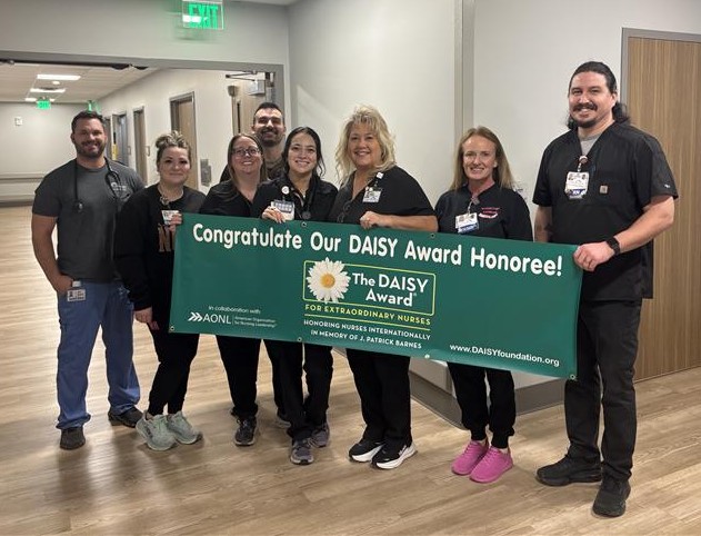 A group of eight smiling healthcare workers stand in a hallway holding a green banner that reads, Congratulate Our DAISY Award Honoree! For Extraordinary Nurses. They are wearing scrubs and name badges.