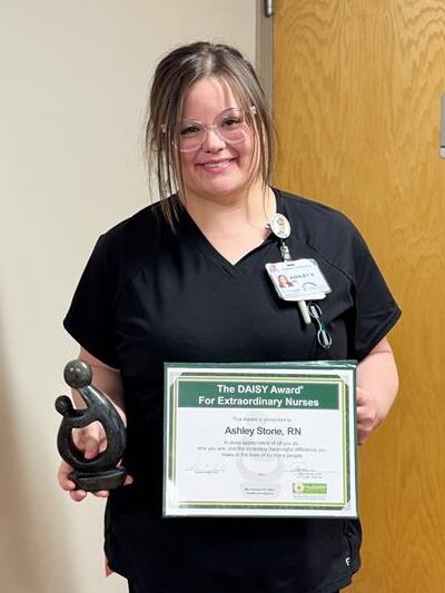 A woman in black scrubs stands indoors, smiling and holding a framed DAISY Award certificate and a small statue. She has glasses and a name badge, with a closed wooden door in the background.