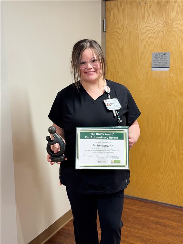 A woman in black scrubs stands indoors, smiling and holding a framed DAISY Award certificate and a small statue. She has glasses and a name badge, with a closed wooden door in the background.