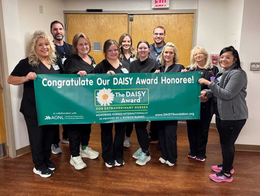 A group of nurses and hospital staff, all in scrubs, smile while holding a green banner that reads “Congratulations Our DAISY Award Honoree!” with the DAISY Foundation logo and website information.