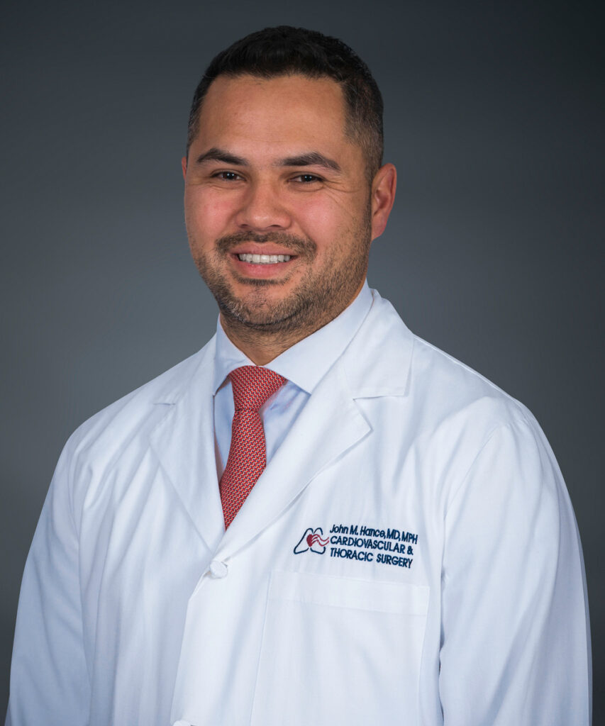 A man with short dark hair and a trimmed beard, wearing a white medical coat that reads Cardiovascular & Thoracic Surgery, smiles against a dark gray background&mdash;highlighting his expertise in minimally invasive heart pump procedures.
