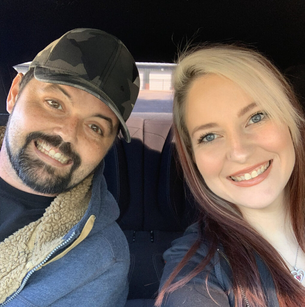 A man with a beard and a camo hat and a woman with long, blonde hair are sitting in a car, smiling at the camera.