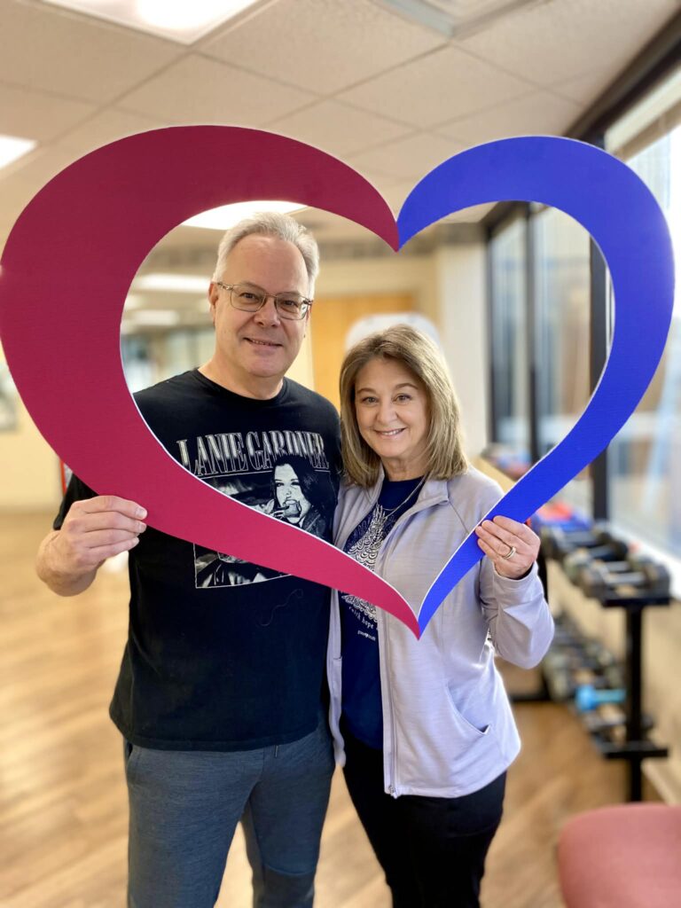 A smiling man and woman stand indoors, holding a large heart-shaped frame together—perfect for celebrating their cardiac rehab journey. The frame is red on one side and blue on the other, while sunlight streams through windows in the background.